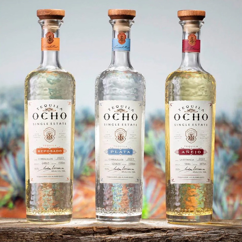 Three bottles of Tequila Ocho on a wooden surface with a blurred agave background.