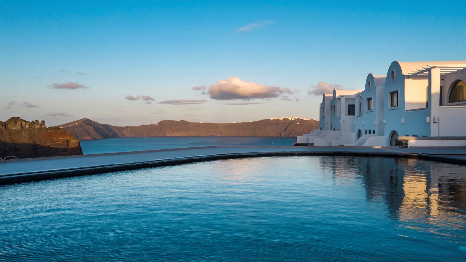 Infinity pool overlooking the Aegean Sea with white buildings and a mountainous coastline in the background during sunrise.