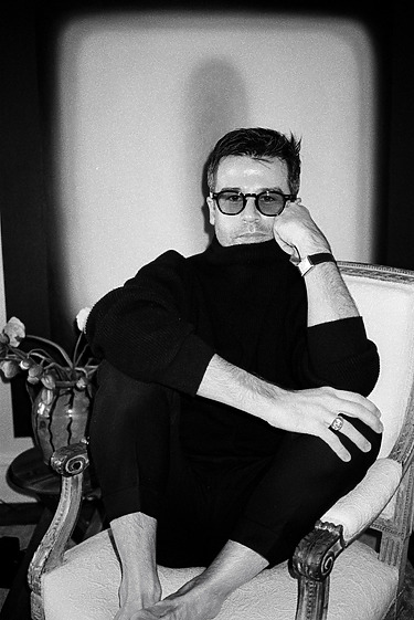man sitting in a chair wearing glasses and a dark sweater, resting head on hand, monochrome photo