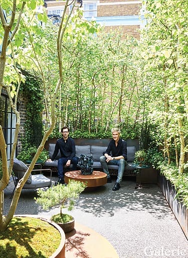 Two people sitting in a lush garden patio surrounded by tall, green plants and trees, with a round wooden table between them.