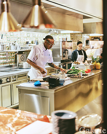 Chef preparing food in a modern kitchen with various ingredients on the counter, while another chef works in the background.