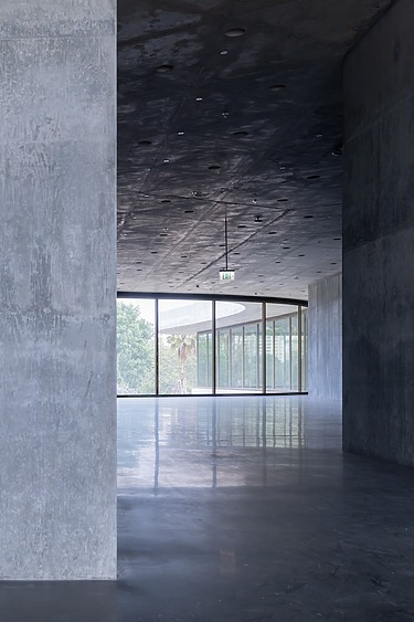 Modern building interior with concrete columns and large windows offering a view of trees outside.