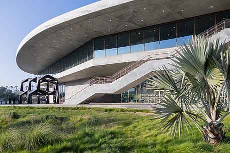 Modern building with curved roof, large windows, and geometric outdoor sculpture, surrounded by grass and a small tree.