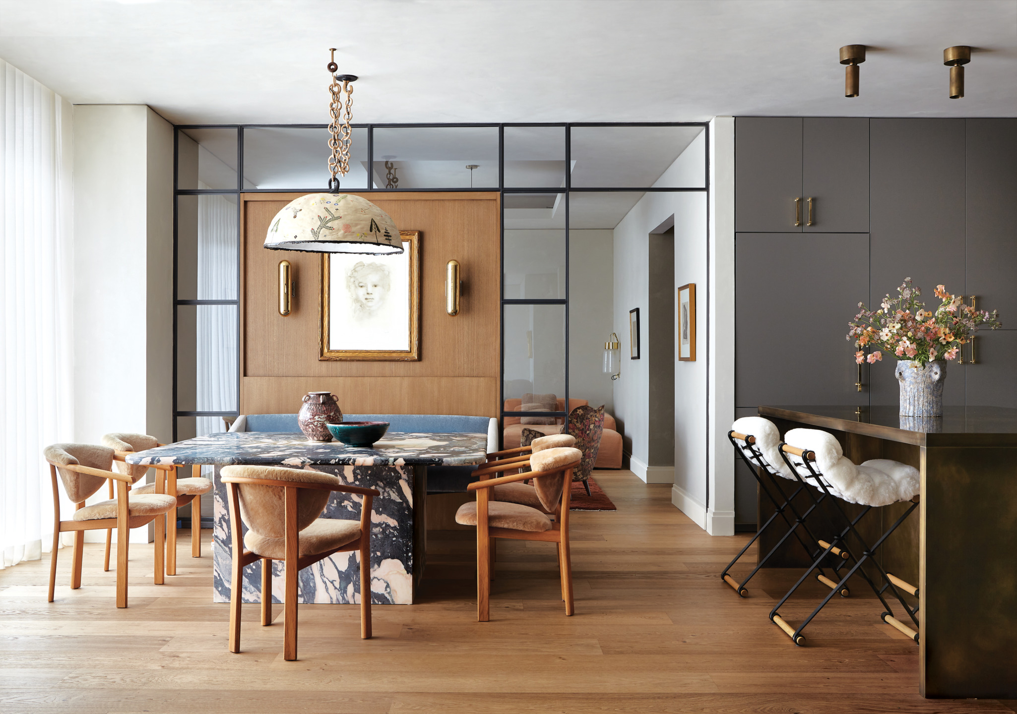 Modern dining room with wooden chairs, marble table, pendant light, floral arrangement on counter, and eclectic decor elements.