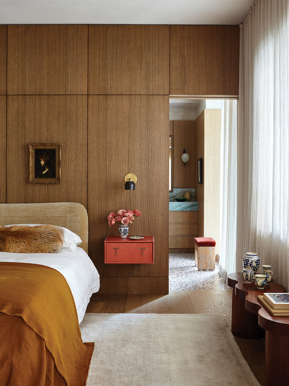 Cozy bedroom with wood paneling, a bed with brown and orange linens, a red nightstand, and a view into an adjoining room.