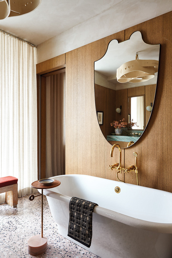 Modern bathroom with freestanding tub, unique mirror, gold fixtures, terrazzo floor, and wooden accent walls.