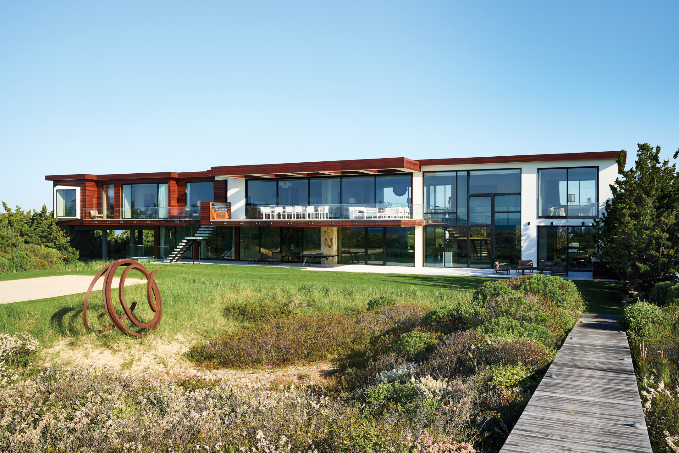 Modern beachfront house with large windows, surrounded by greenery, under a clear blue sky.