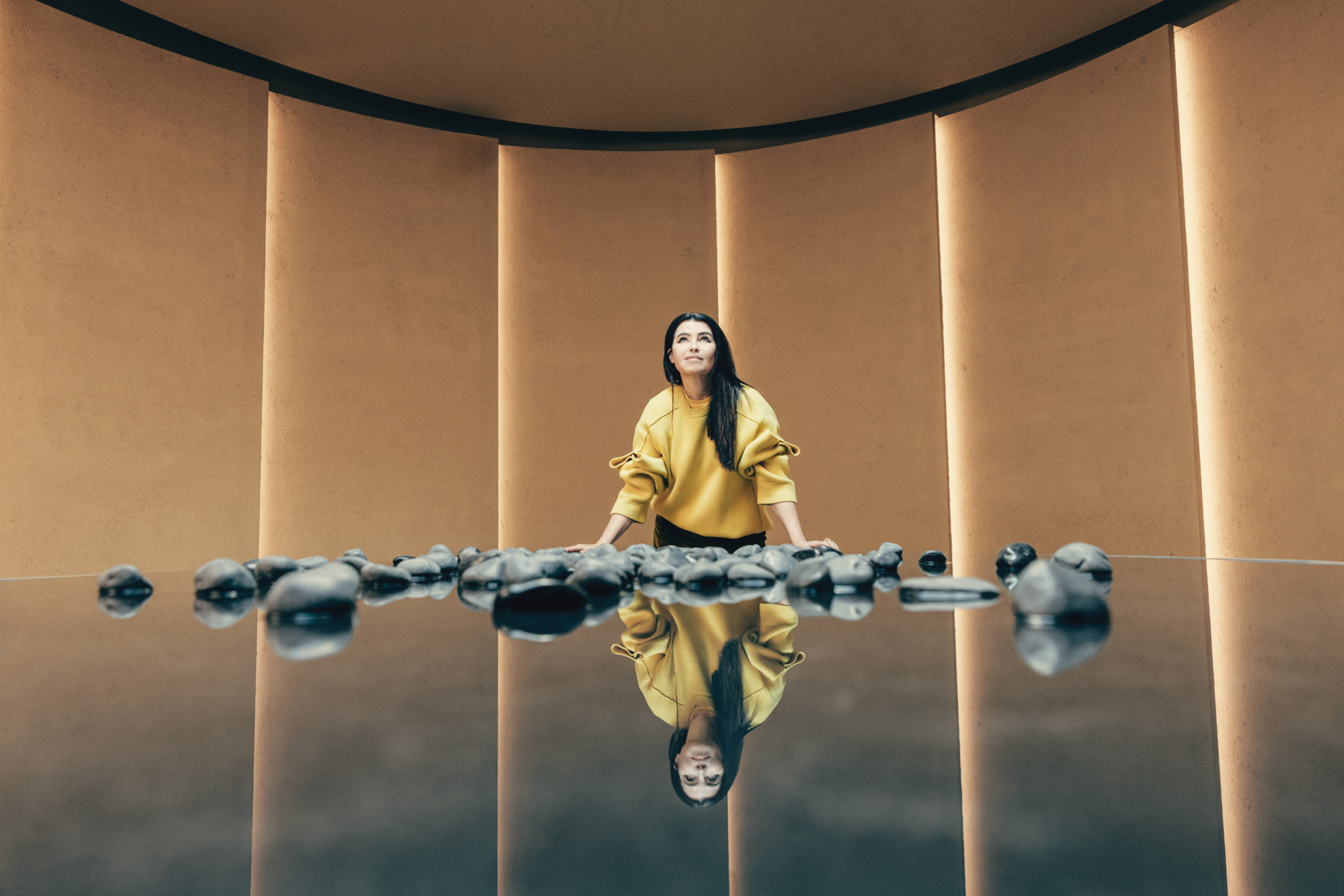 Woman in a yellow sweater kneeling on a reflective surface with stones, surrounded by warm-toned vertical panels.