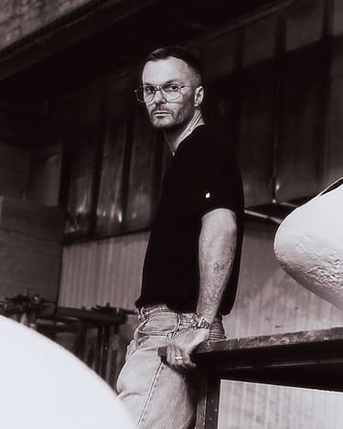 Man in black shirt and jeans leaning against industrial railing, wearing glasses, with a thoughtful expression.