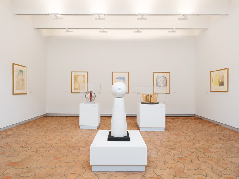 Art gallery room with abstract sculptures and framed artworks on white walls, lit by modern ceiling lights.