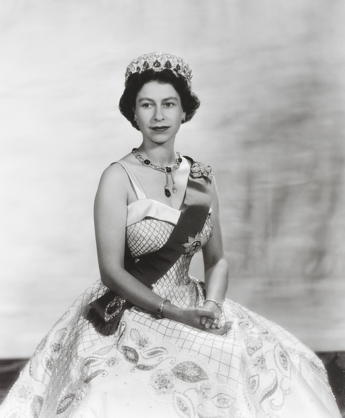 A formal portrait of a woman wearing a crown and sash with a jeweled necklace and elegant gown against a neutral background.