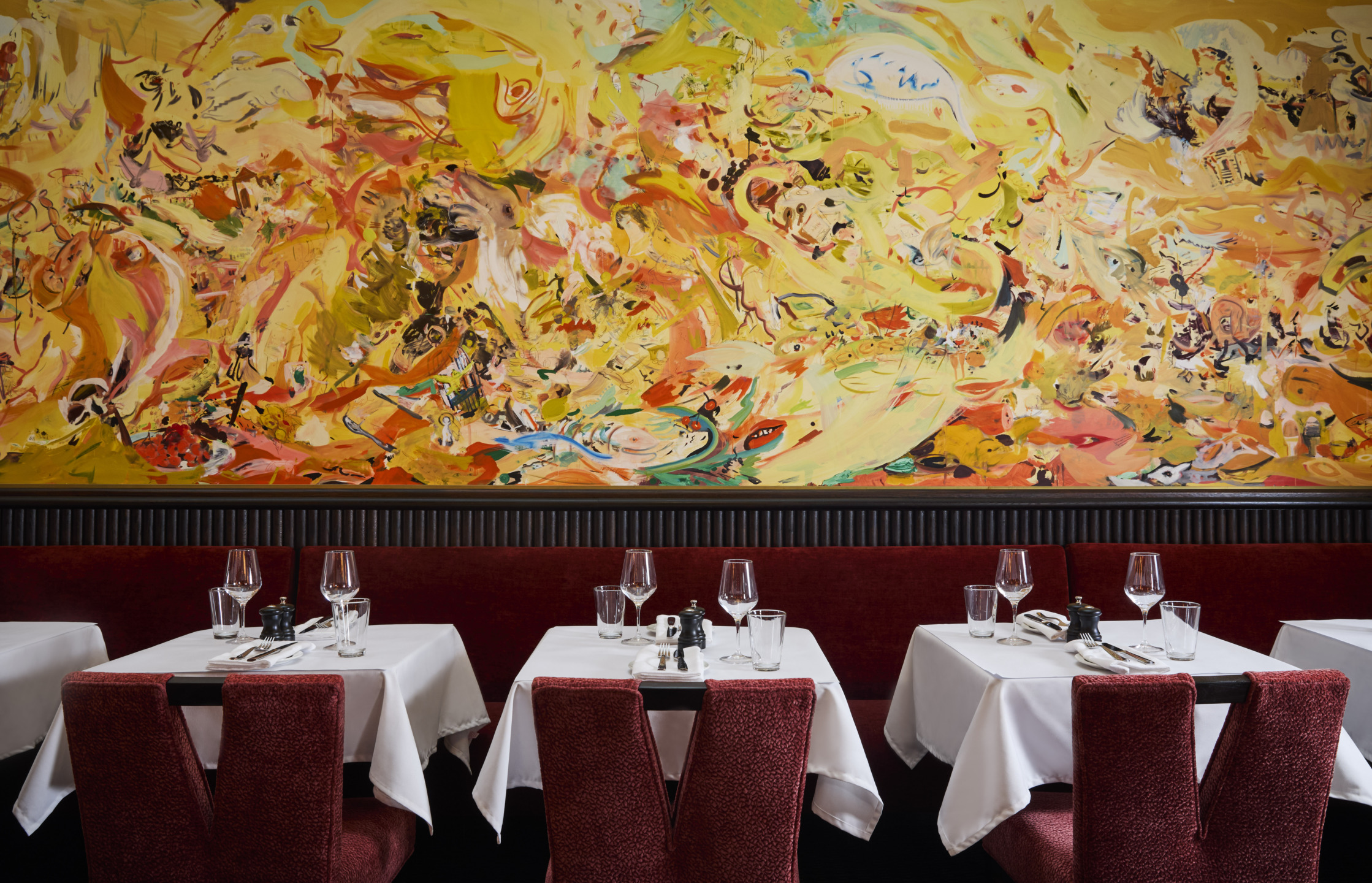 Colorful abstract mural above elegantly set tables with red chairs in a restaurant dining area.