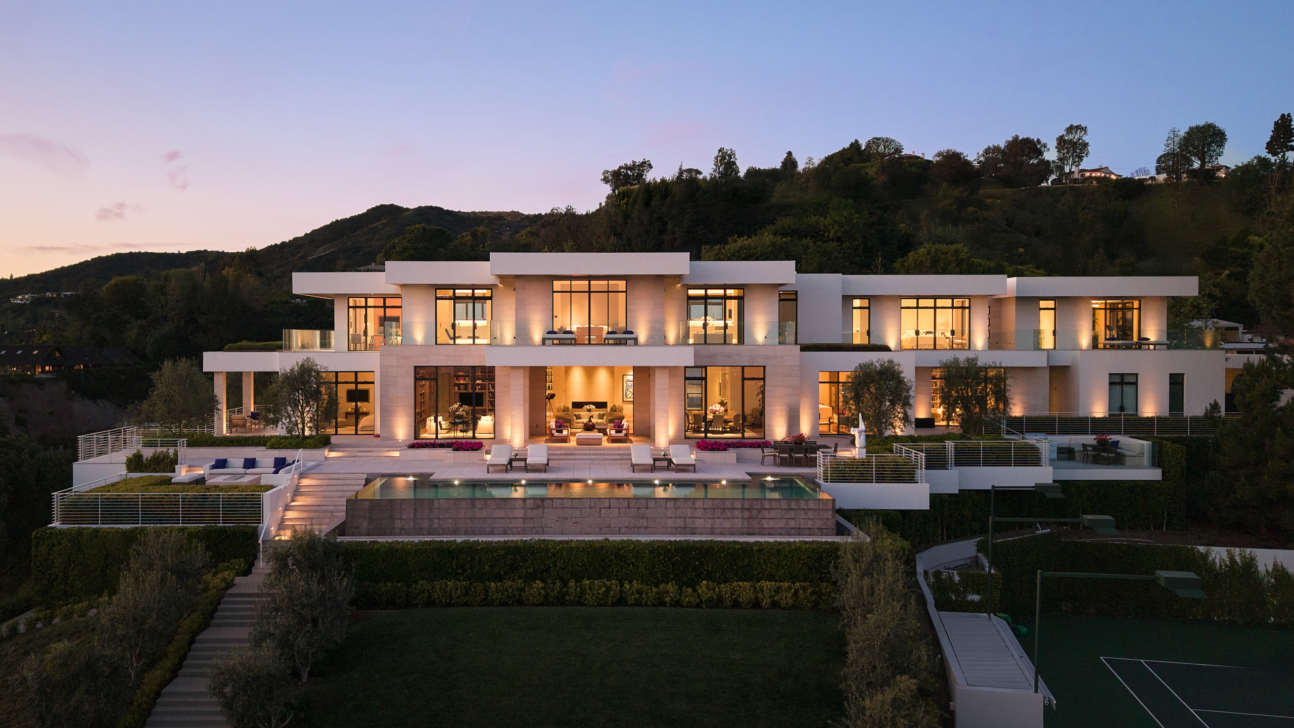 Modern luxury mansion with large glass windows, terrace, pool, and surrounding greenery against a twilight sky.