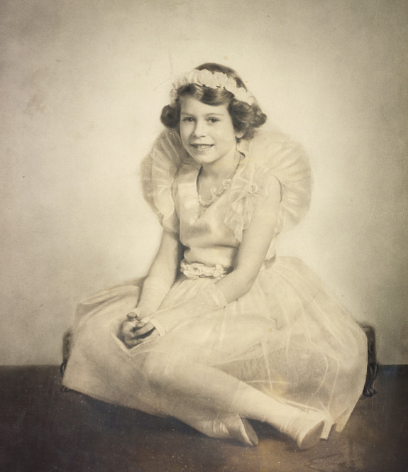 Young girl in vintage dress and crown, sitting and smiling with hands clasped, sepia tone.