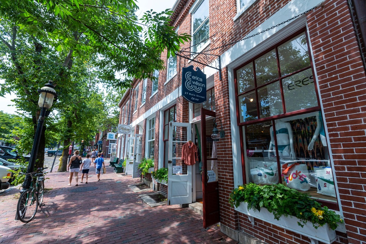 Charming street with red brick buildings, lush green trees, and people walking on a sunny day.
