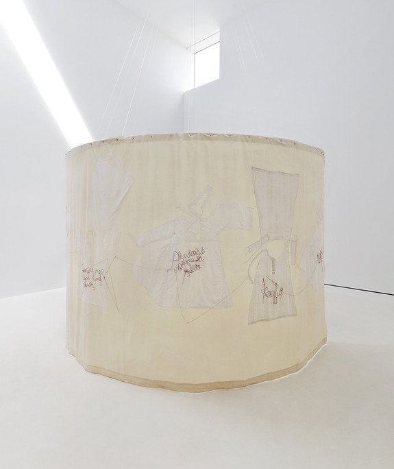 Art installation featuring a circular beige fabric with embroidered designs and text, displayed in a minimalist gallery setting.
