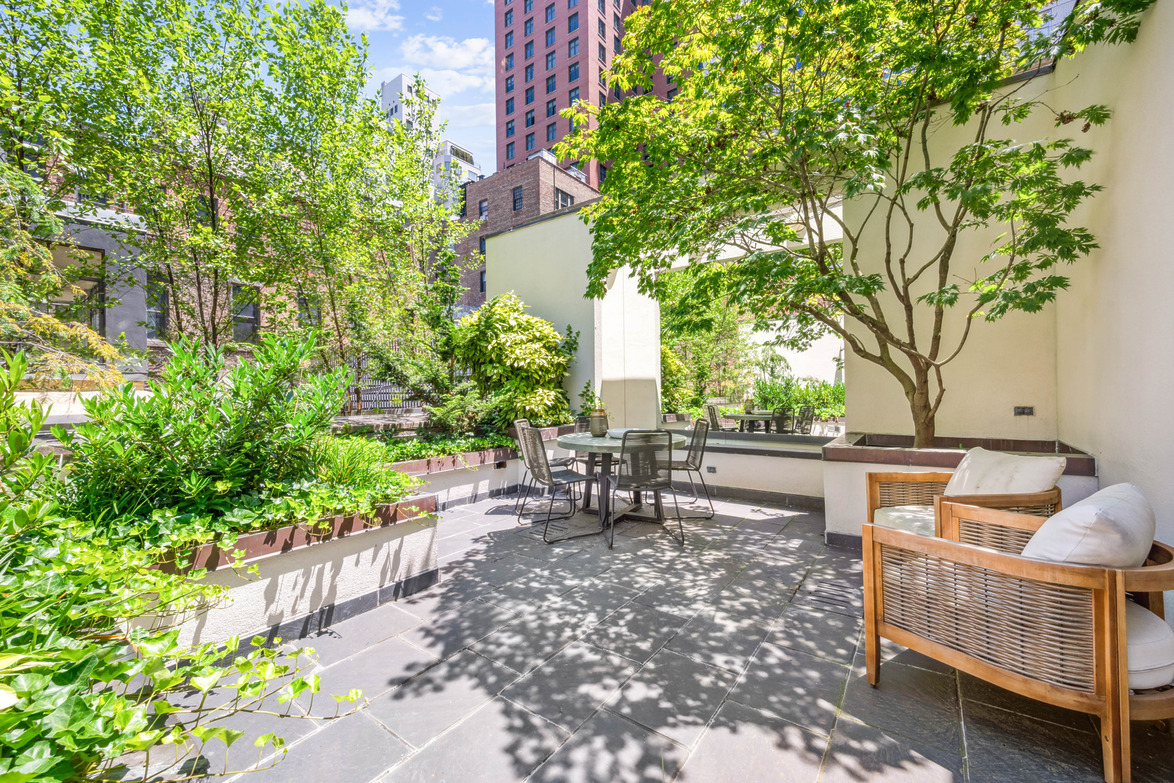 Rooftop garden terrace with greenery, seating area, and city buildings in the background under a clear blue sky.