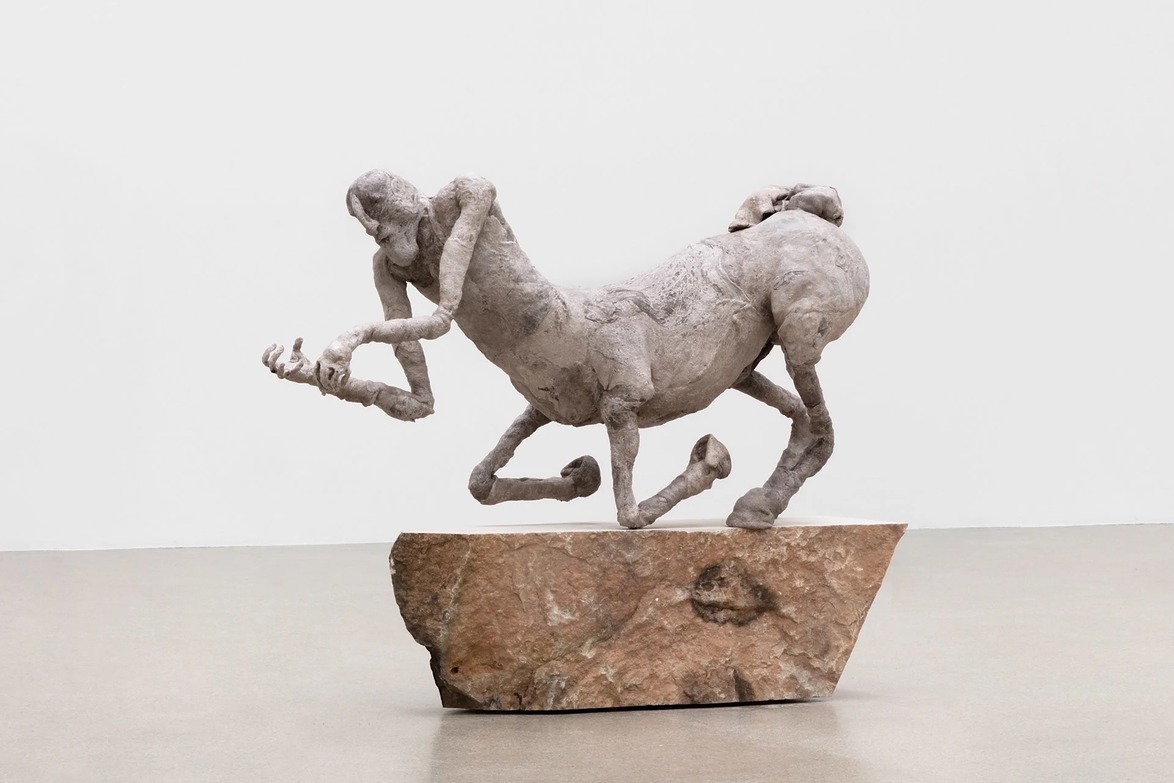 Stone sculpture of a centaur-like creature balancing on a rock, featured in a minimalistic gallery setting.