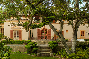 Mediterranean-style home with large arched windows and lush garden, surrounded by mature trees and greenery.