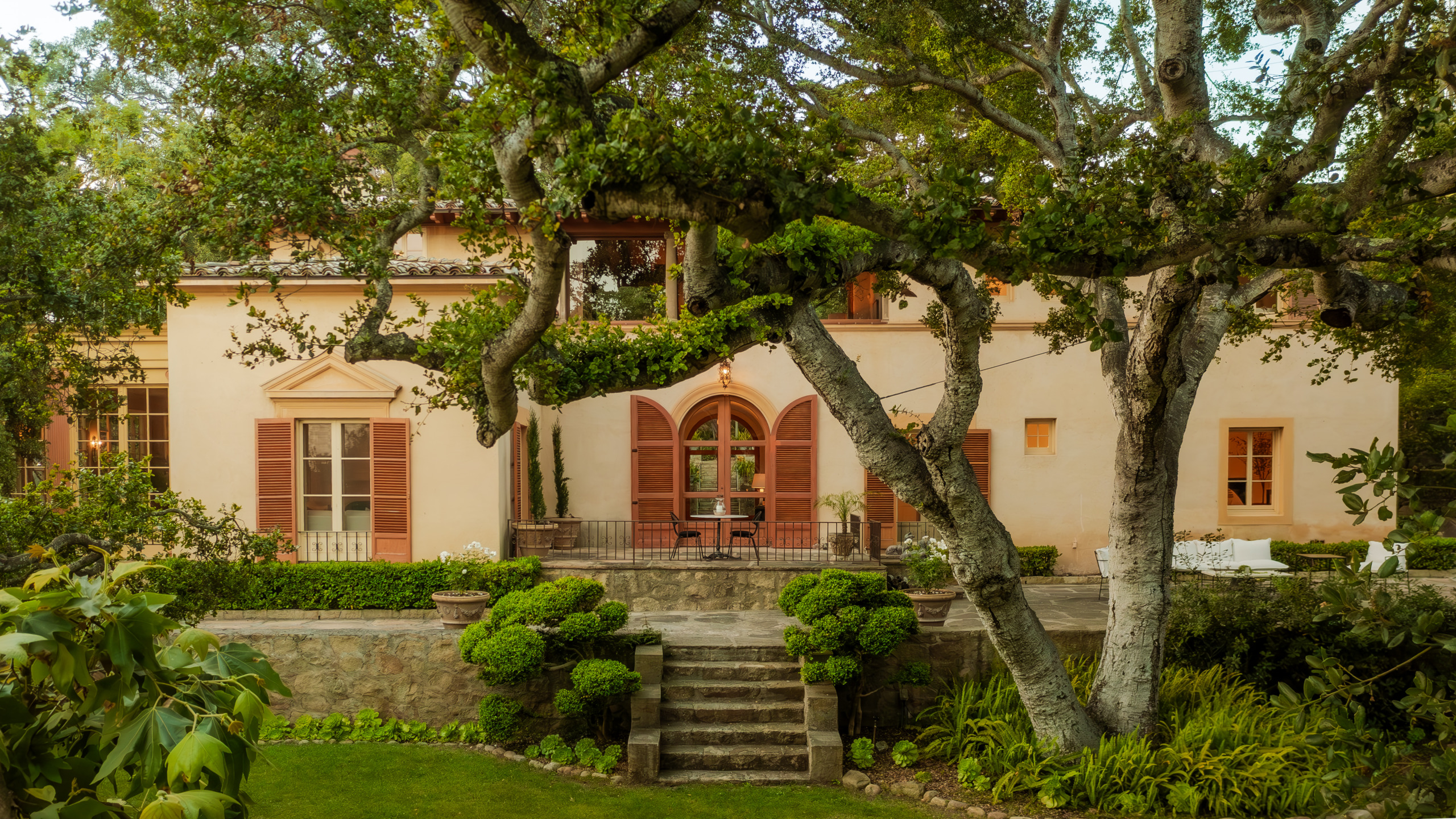 Mediterranean-style home with large arched windows and lush garden, surrounded by mature trees and greenery.