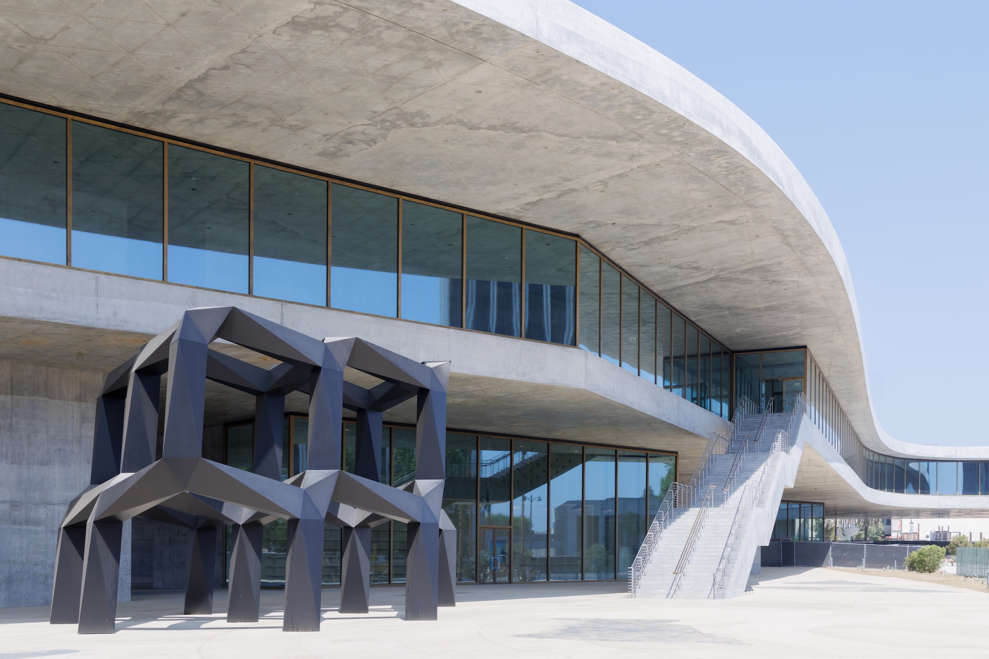 Modern building with glass facade and large abstract sculpture outside, featuring curving architectural lines.