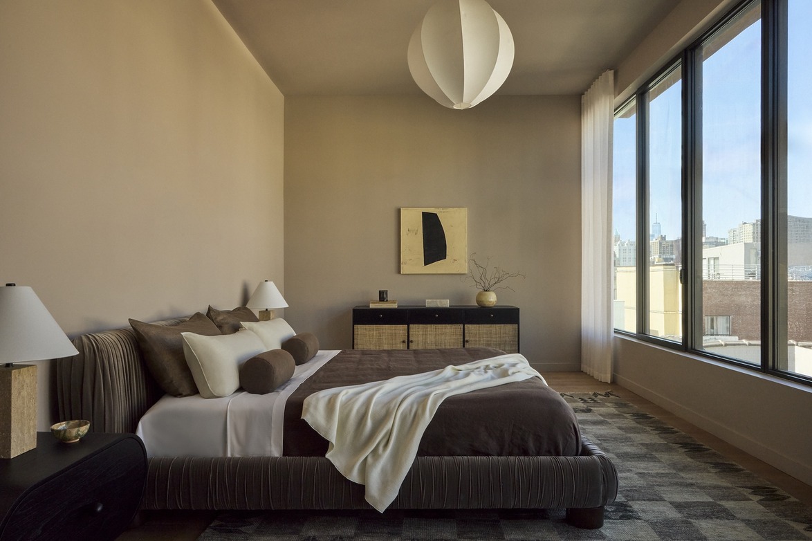 Modern bedroom with large windows, bed with brown and white linens, dresser, decorative pendant light, and wall art.