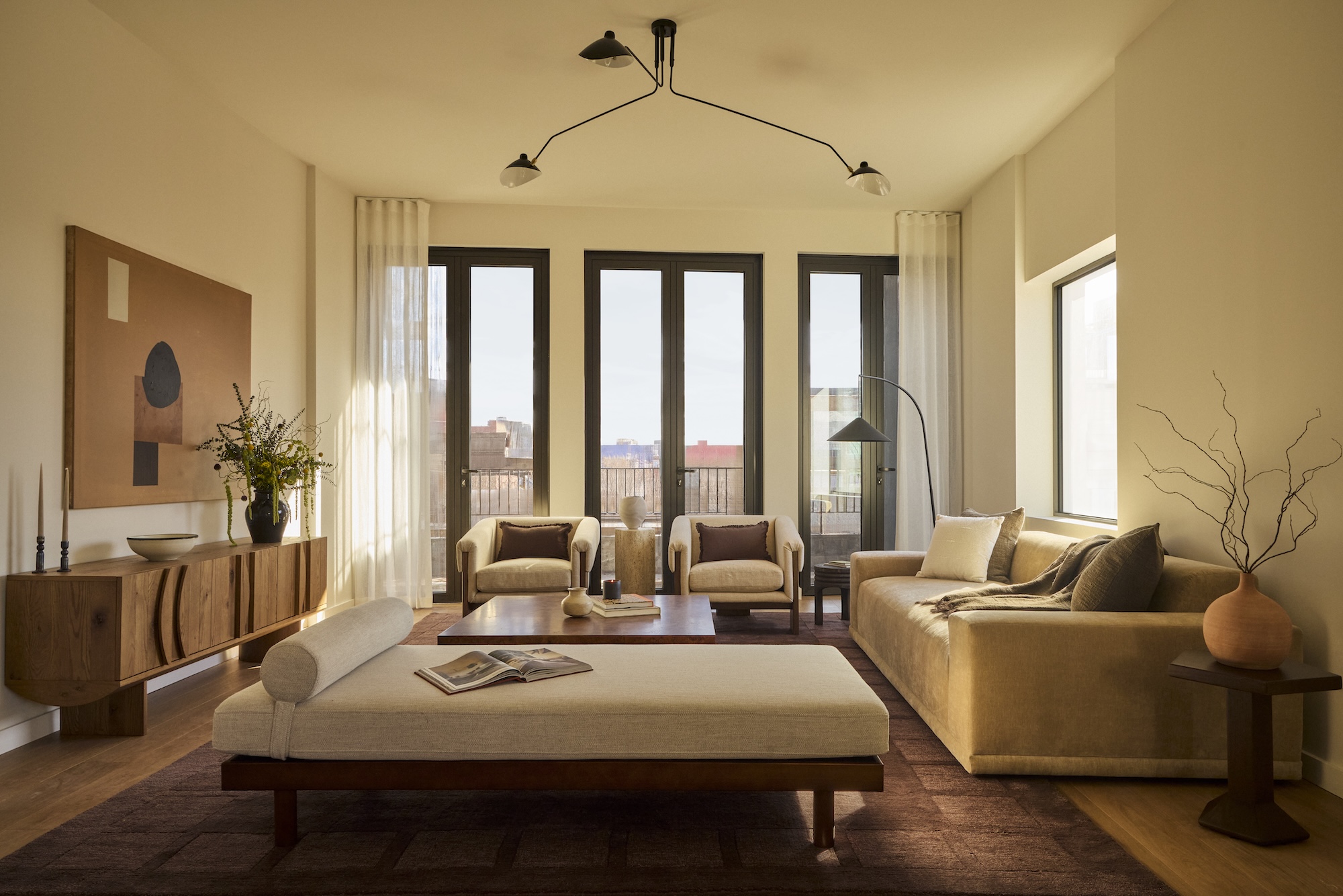 Modern living room with beige furniture, large windows, a wooden sideboard, and decorative plants.