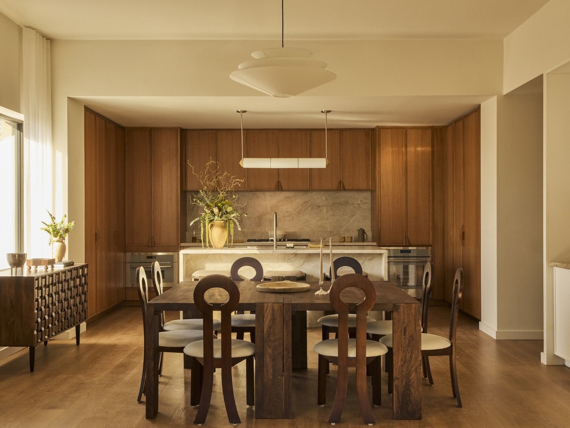 Modern kitchen with wooden cabinets, dining table, decorative plants, pendant lights, and natural light from large window.