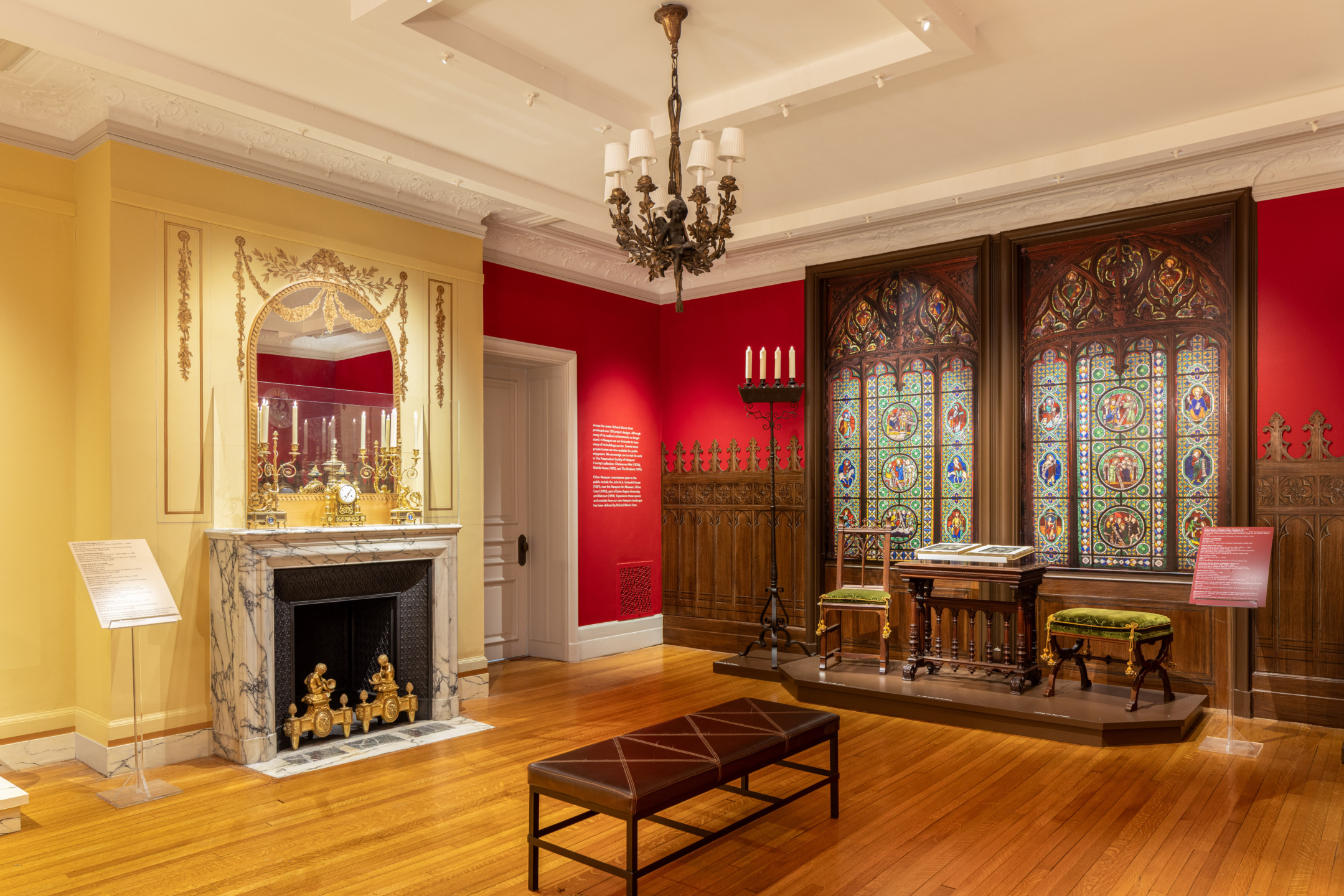 Museum room displaying a fireplace, ornate mirror, stained glass windows, wooden desk, chairs, and red and yellow walls.