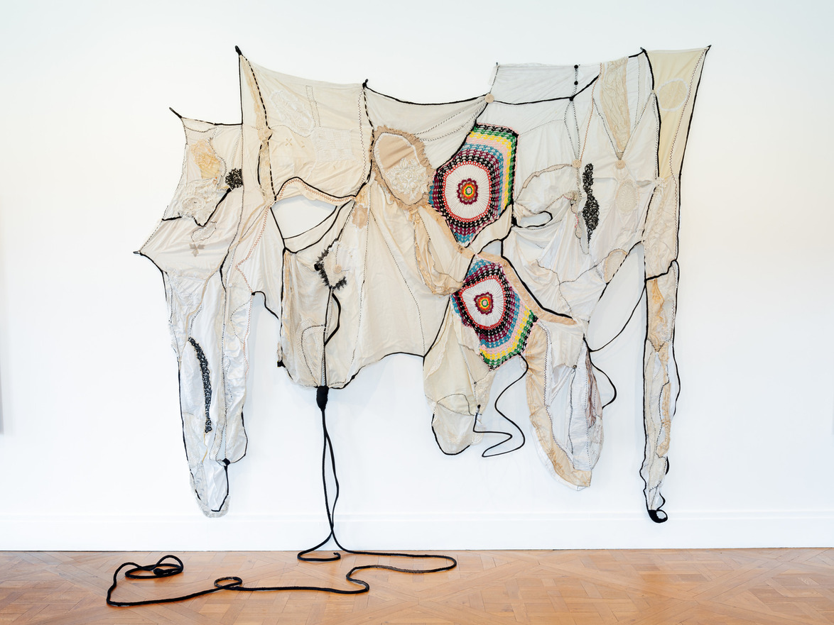 Abstract textile artwork hanging on a white wall, featuring multicolored circular patterns and black cords.