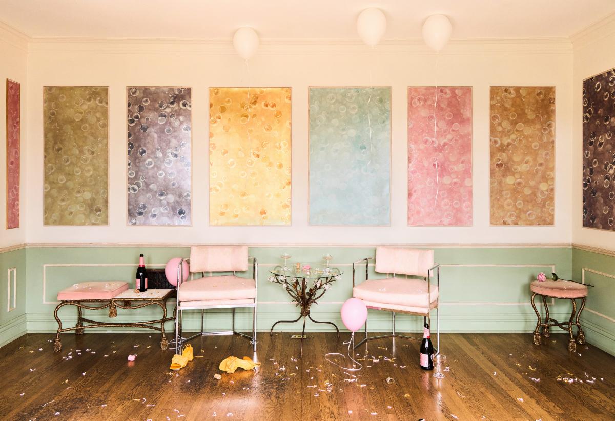 Elegant party aftermath with balloons, champagne bottles, and chairs in a pastel-colored room with abstract art on the walls.