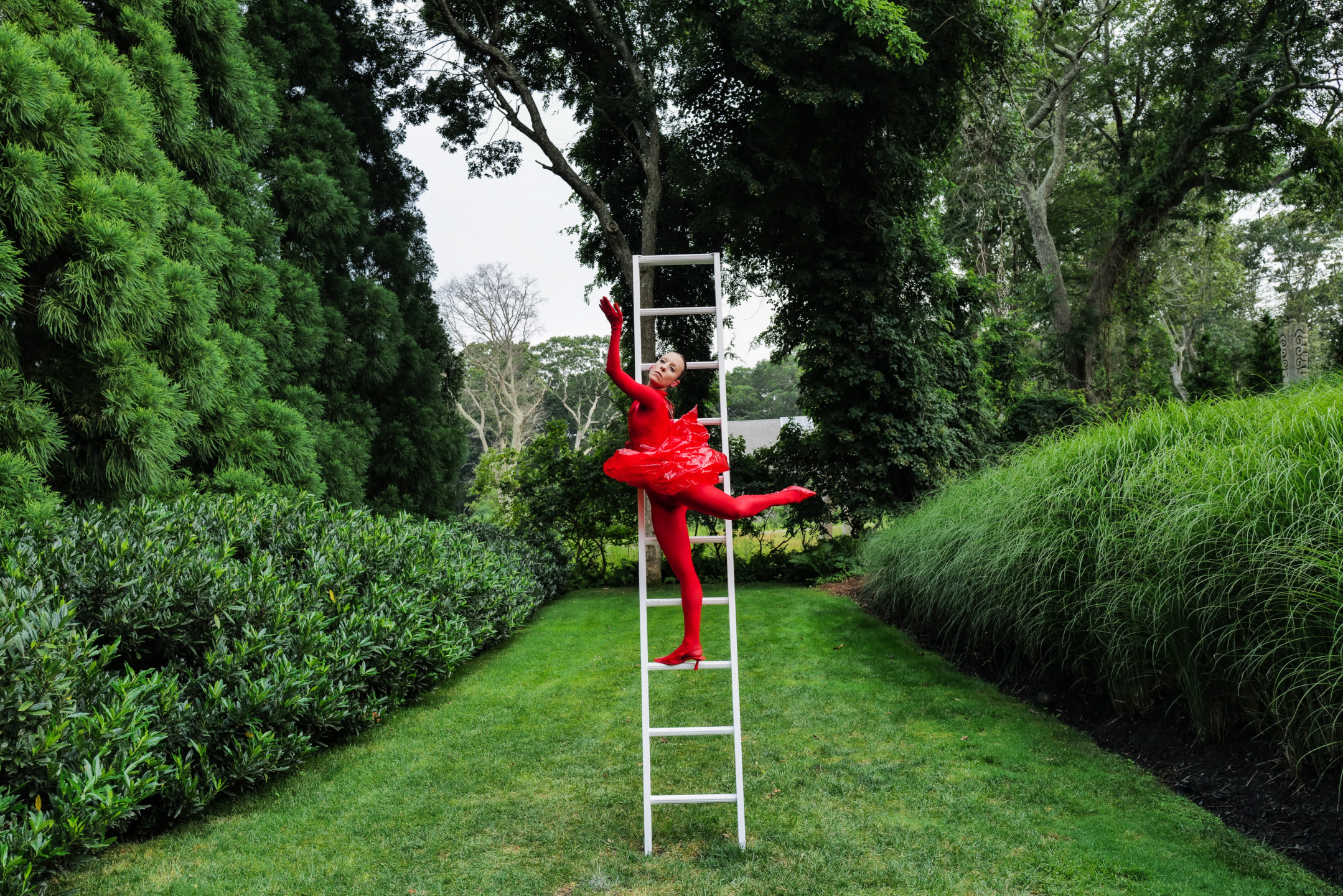 Dancer in a bright red costume poses on a tall white ladder surrounded by lush green trees and grass in a garden.
