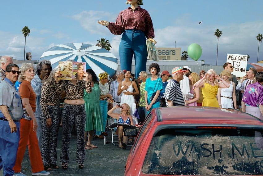 Surreal scene with a crowd, oversized woman figure, and a dusty car with "Wash Me" written on it, set in a sunny urban area.