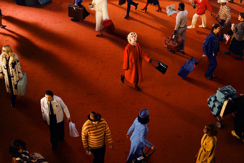 People walking on a red carpeted floor, carrying suitcases and bags, wearing colorful coats and hats, from a top view.