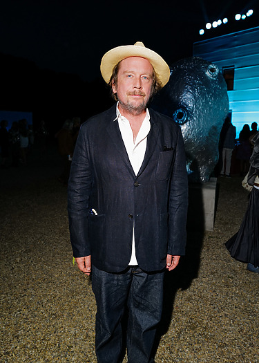 Man in dark suit and straw hat stands outdoors at evening event with large sculpture and lit background.