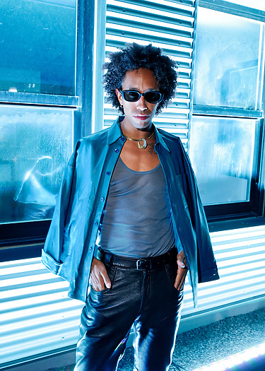 Person wearing sunglasses and stylish outfit stands confidently against a metallic background with blue lighting effects.