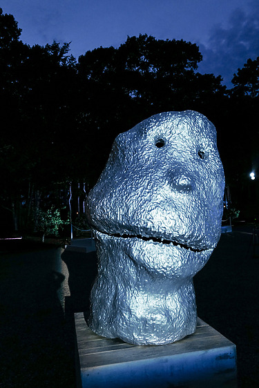 Sculpture of a large metallic head with abstract features set outdoors against a dark evening backdrop.