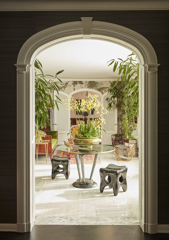 Elegant indoor garden room with plants, stools, and a central table with a large bowl of yellow orchids under an arched doorway.