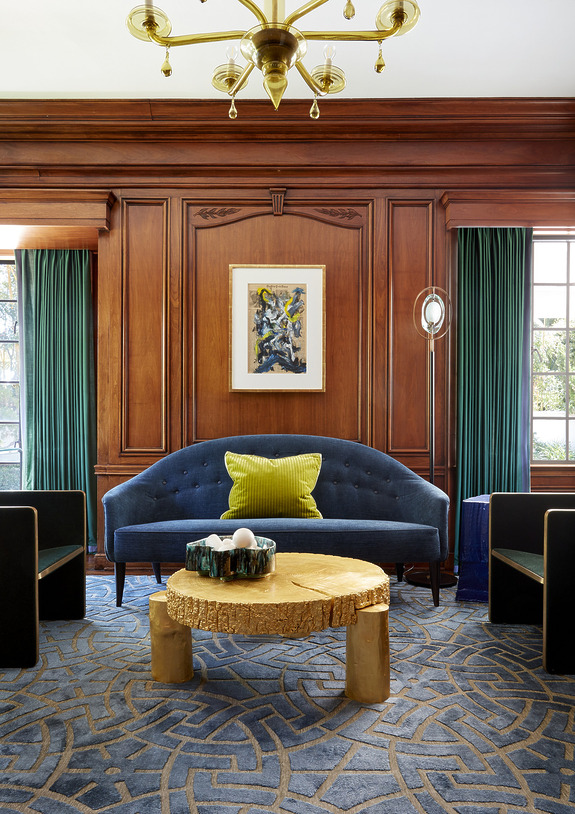 Luxurious living room with blue sofa, wooden paneling, abstract painting, geometric rug, and unique wooden coffee table.