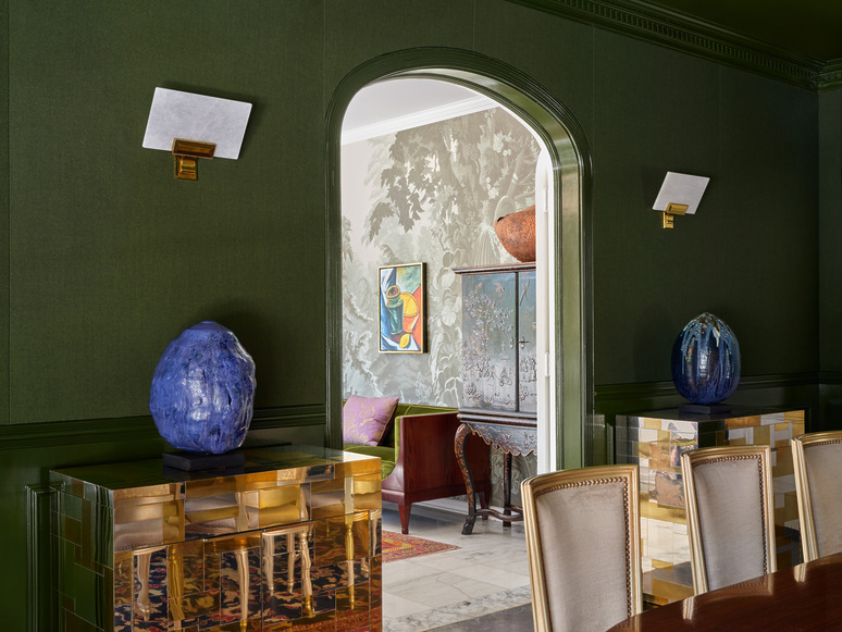 Elegant dining room with green walls, modern art, and stylish decor, featuring sculptural elements and a glimpse into an adjoining room.