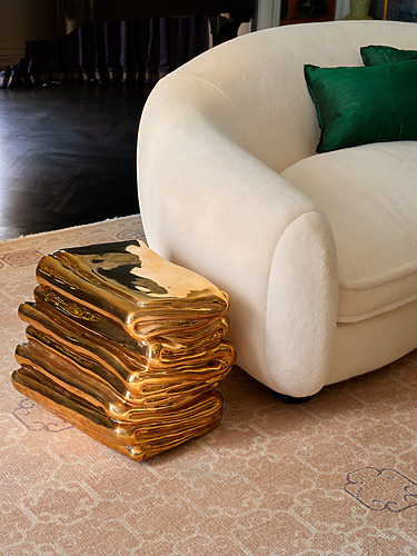 Modern living room with a beige couch, green cushions, and a unique gold stacked book-shaped side table on a patterned rug.