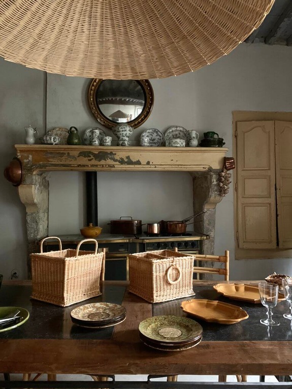 Rustic kitchen with woven baskets on a wooden table, decorative plates, and vintage pots on a stone mantelpiece.