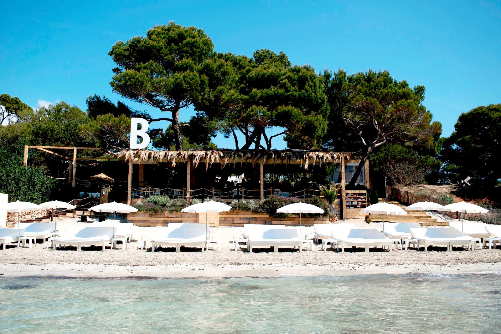 Beachfront with white loungers and umbrellas, wooden beach bar surrounded by trees in the background, clear blue sky overhead.