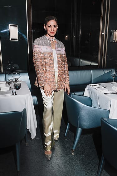 Person wearing a colorful jacket and satin pants standing in a stylish dining area with set tables and teal chairs.