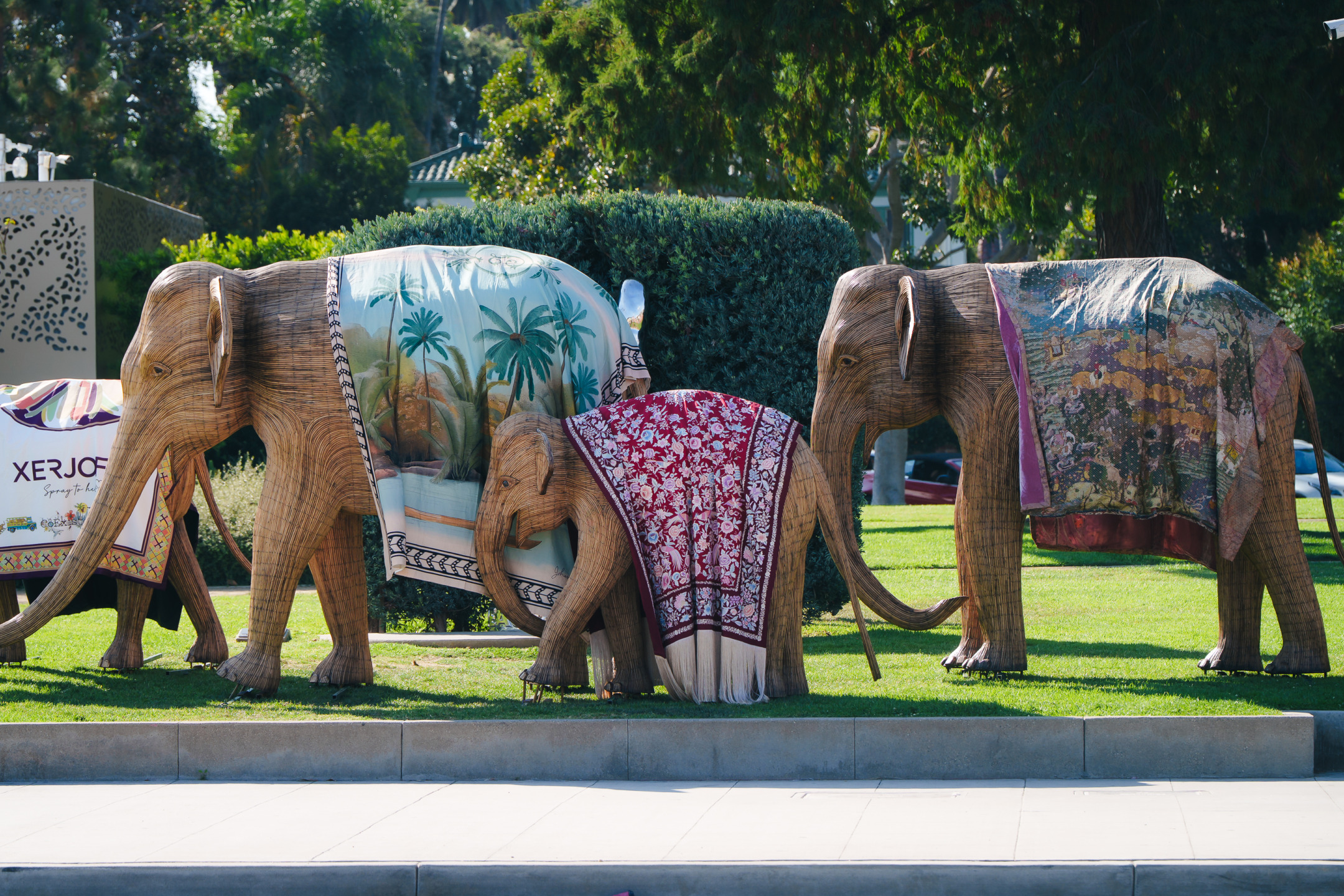 Three wooden elephant sculptures draped in colorful fabrics, standing on grass with trees in the background.