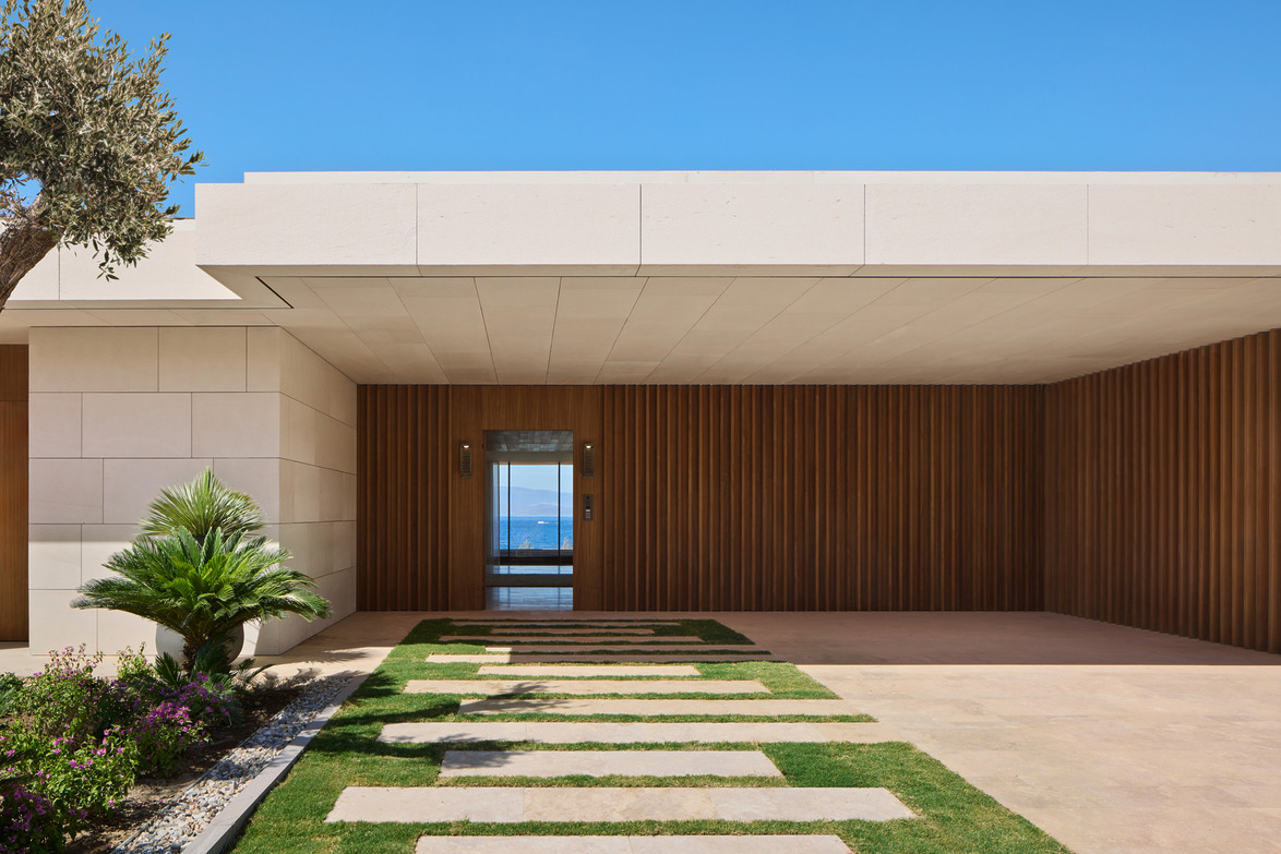 Modern house entrance with wooden facade, glass door, and stone walkway surrounded by green grass and plants.