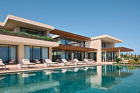 Luxurious modern villa with large pool, surrounded by lounge chairs and greenery under a clear blue sky.