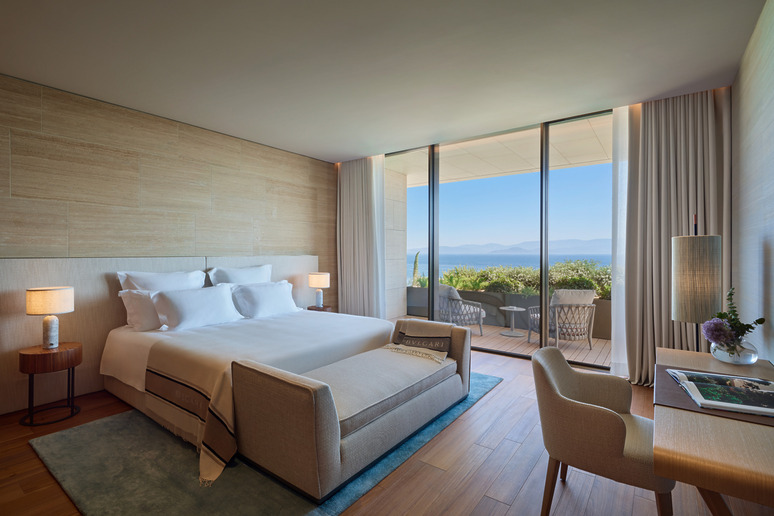 Modern hotel room with a large bed, desk, and balcony view of a scenic lake and mountains.
