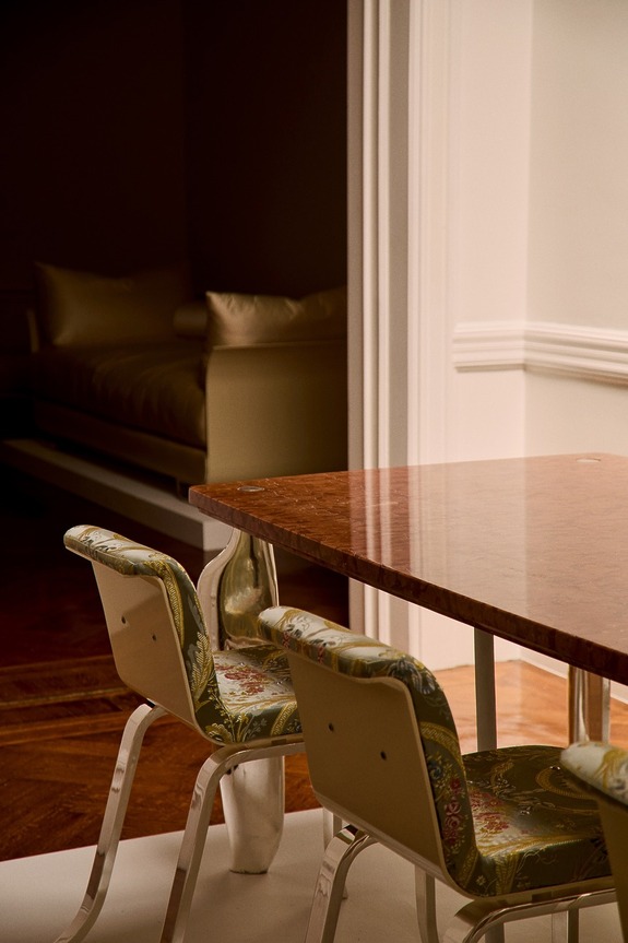 Elegant dining table with two patterned chairs and a glimpse of a luxurious gold sofa in a dimly lit room.