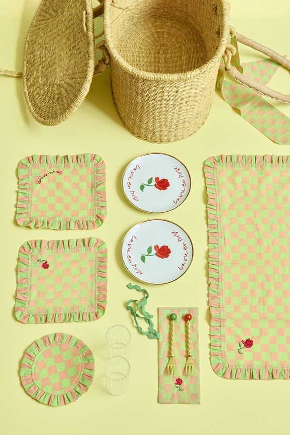 Picnic basket set with rose-patterned plates, green-pink checkered napkins, tablecloth, glasses, and cutlery on yellow background.